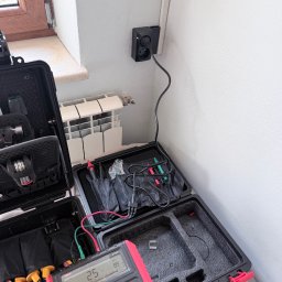 RM Technology - Otwarta walizka z multimetrem i przewodami pomiarowymi podłączonymi do gniazdka elektrycznego. Widoczny fragment okna i grzejnika. Pomiar rezystancji.