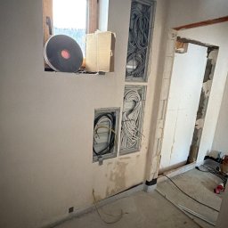 RM Technology - Wnęki w ścianie z poprowadzonymi kablami elektrycznymi. Widoczne kable w peszlach i skrzynkach. Surowy stan wykończenia wnętrza, okno z parapetem.