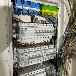 RM Technology - Rozdzielnica elektryczna z zainstalowanymi bezpiecznikami i podłączonymi przewodami. Widoczne oznaczenia obwodów: 'Kuchnia', 'Salon'.