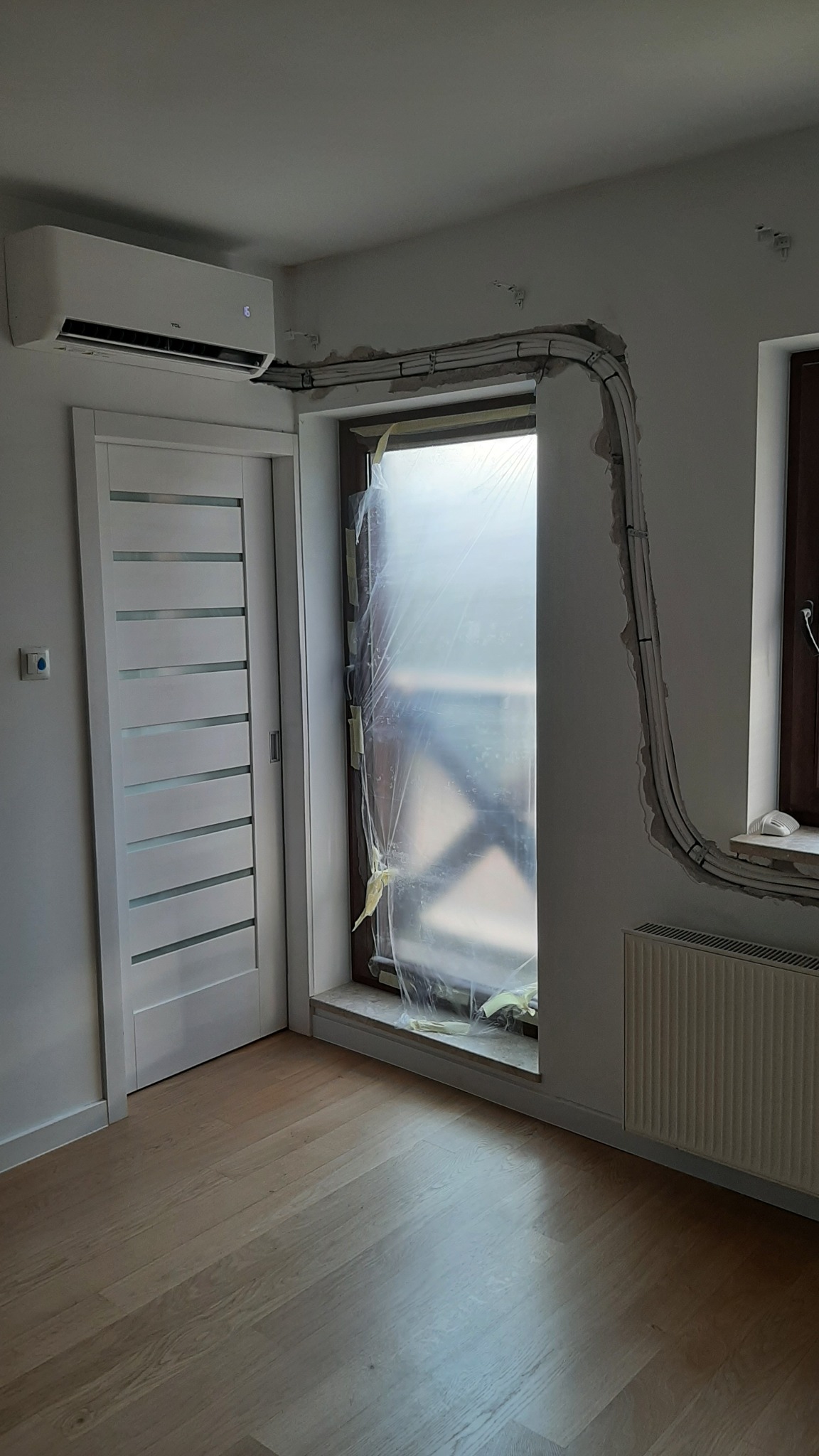 Wnętrze pokoju w trakcie montażu klimatyzacji: widoczny biały klimatyzator ścienny TCL, rury instalacyjne prowadzone wzdłuż ściany nad oknem zabezpieczonym folią, białe drzwi z przeszkleniami...