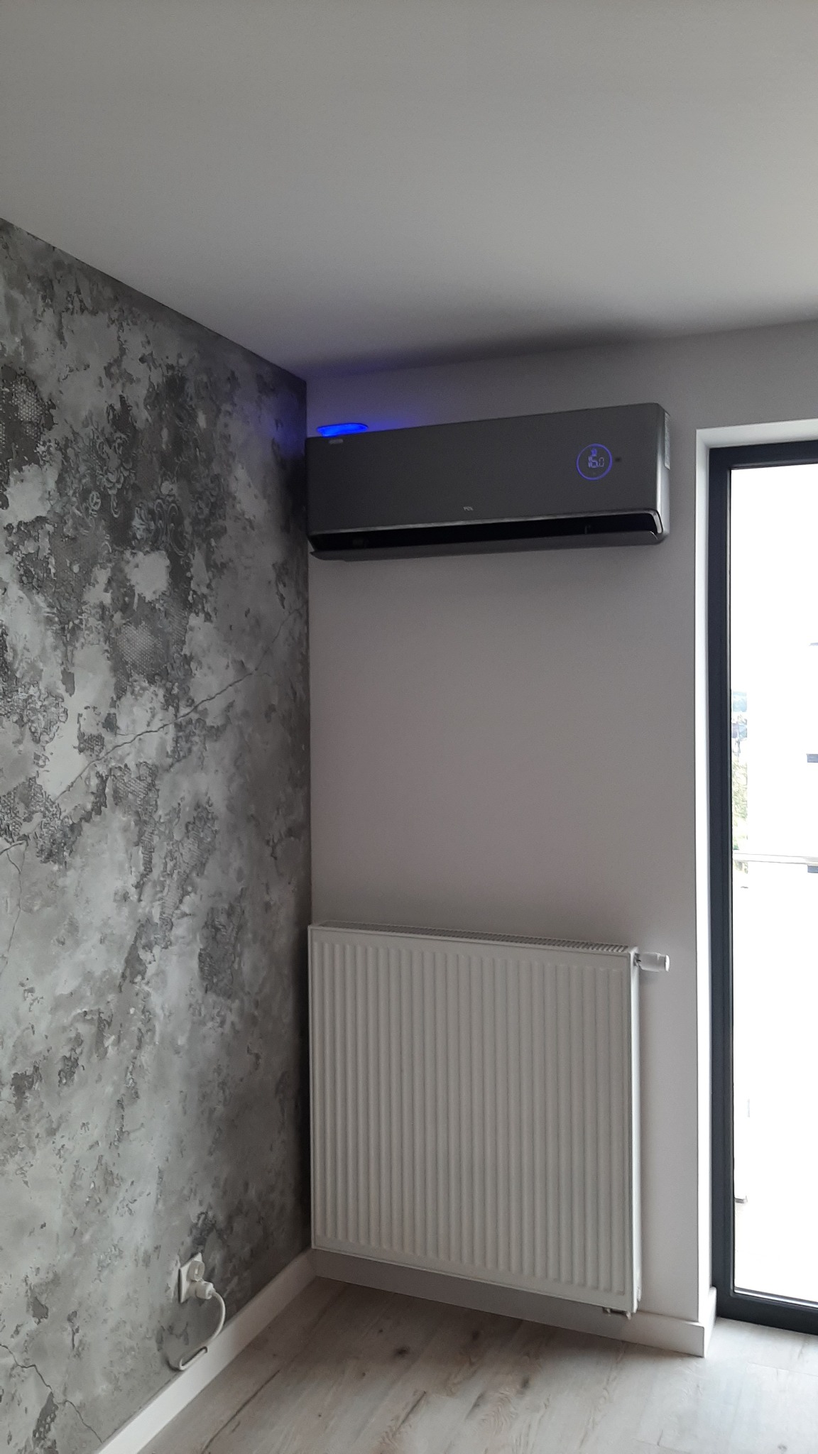Szary klimatyzator TCL zamontowany na ścianie nad białym grzejnikiem w rogu pokoju z dekoracyjną ścianą imitującą beton.
