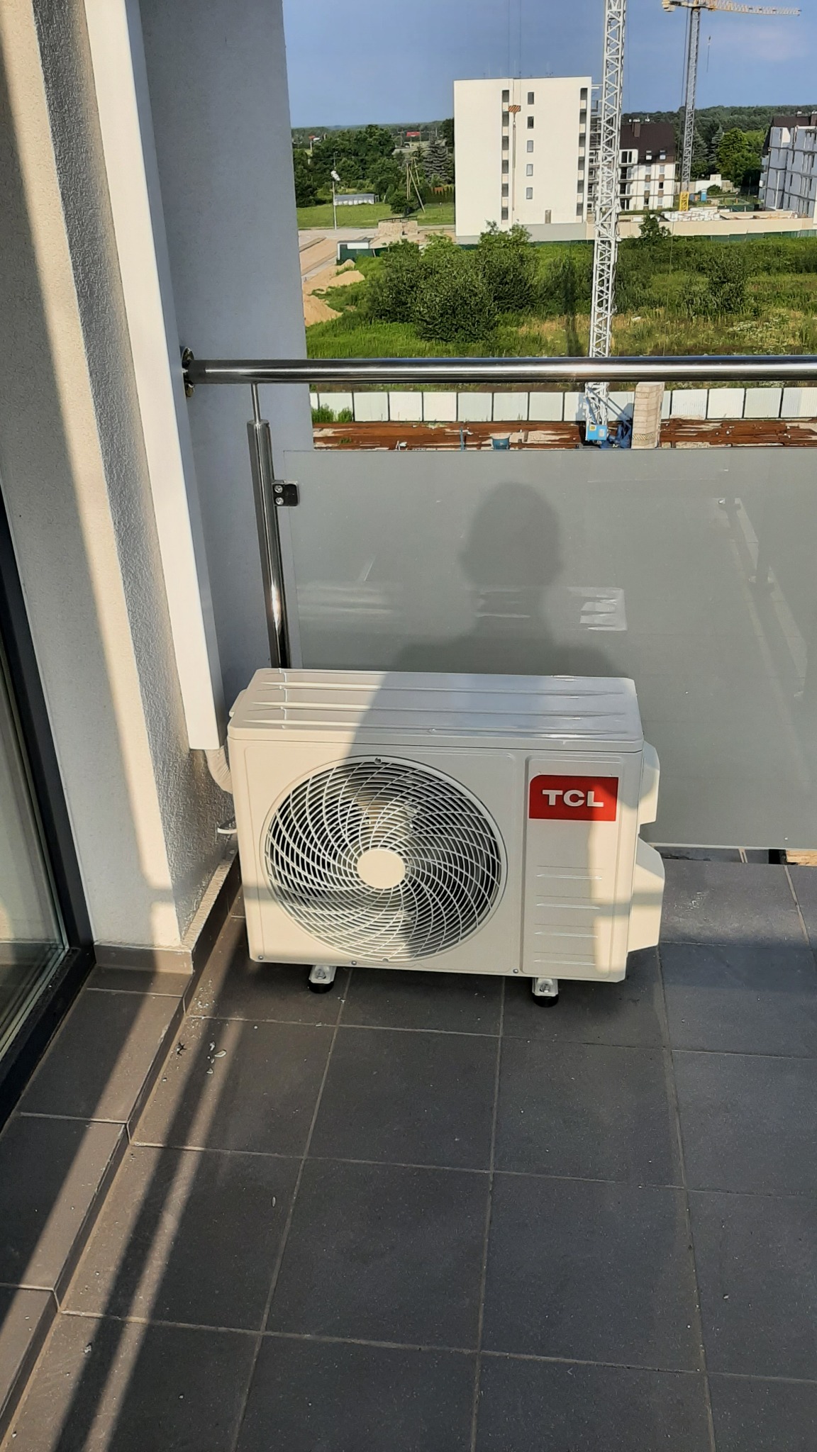 Biała jednostka zewnętrzna klimatyzacji marki TCL na balkonie z ciemnymi płytkami, w tle budynek w budowie i żurawie.