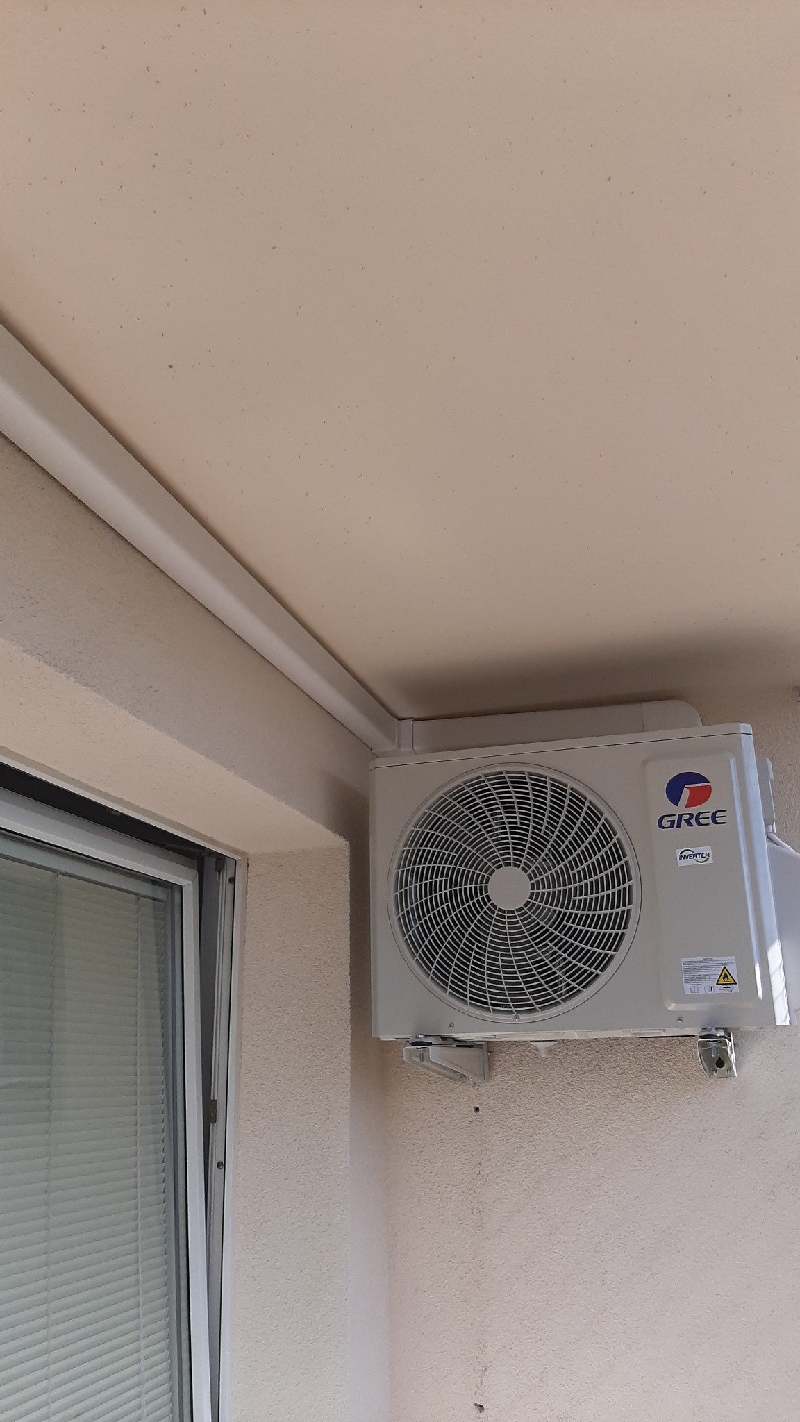 Zewnętrzna jednostka klimatyzacji marki Gree zamontowana na ścianie budynku, widoczna obok okna z żaluzjami.