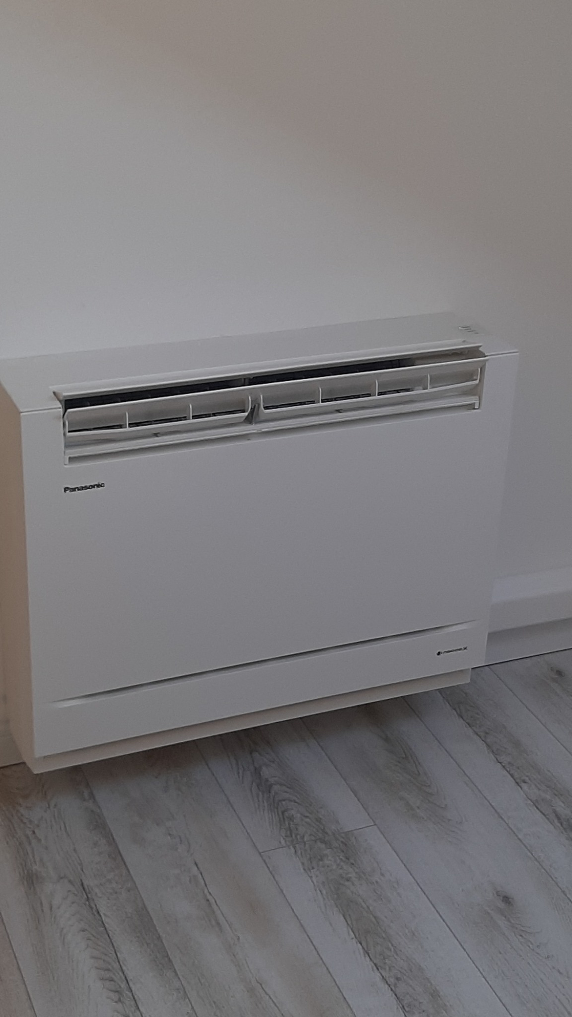 Biały klimatyzator podłogowy Panasonic zamontowany przy ścianie na tle paneli podłogowych imitujących drewno.