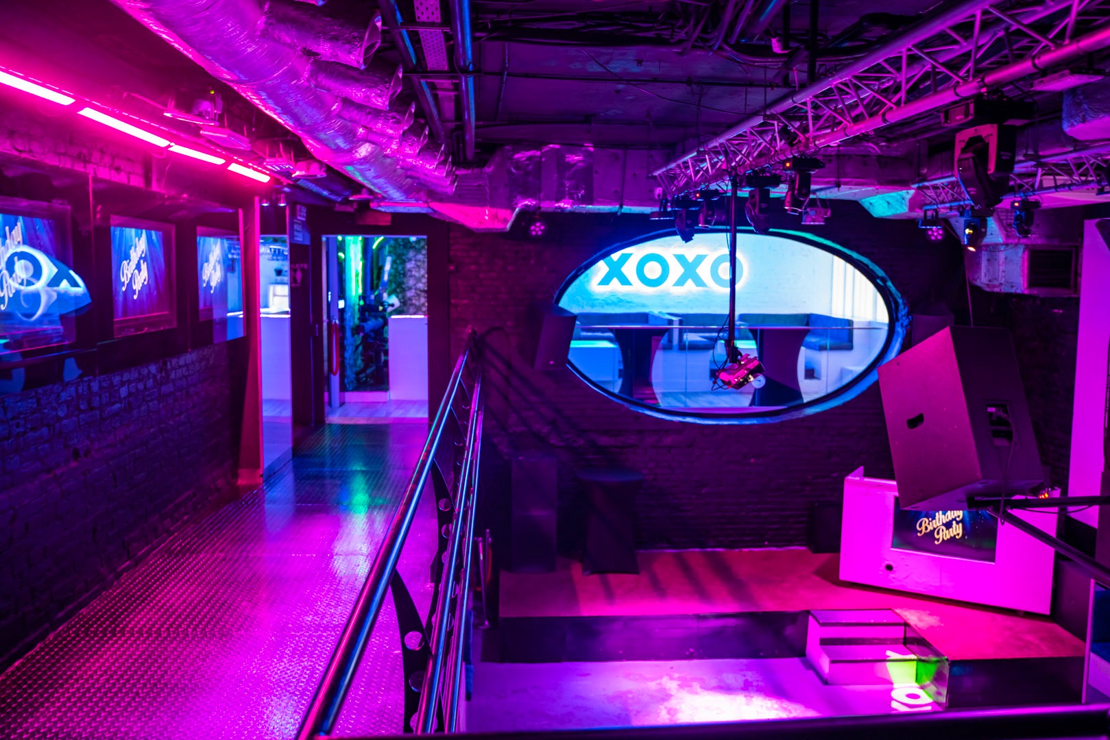 Wnętrze klubu nocnego z neonowym oświetleniem w odcieniach różu i fioletu, widoczna scena z napisem 'Birthday Party' i logo 'XOXO' w okrągłym oknie.
