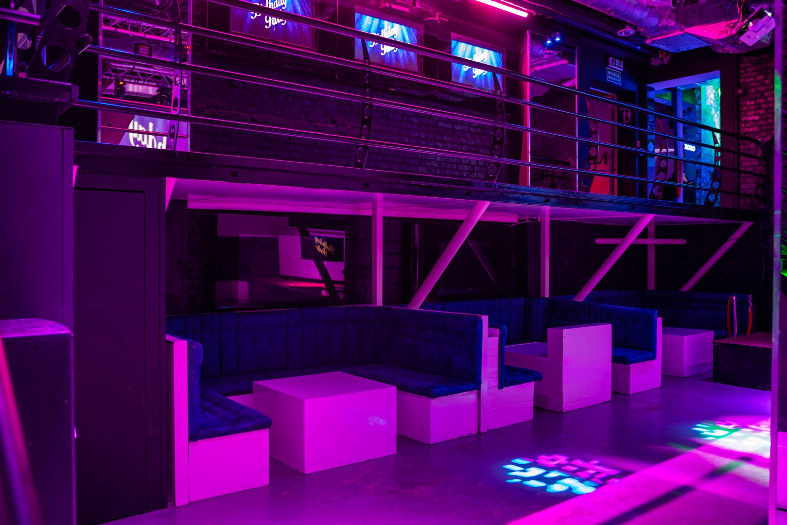 Wnętrze klubu nocnego oświetlone fioletowym i niebieskim światłem, z lożami z granatowymi siedziskami i białymi stolikami, widoczna antresola z monitorami.