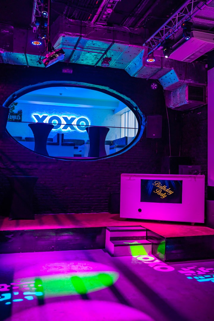 Wnętrze klubu oświetlone neonami w odcieniach różu i fioletu, z dekoracją 'XOXO' widoczną przez owalne okno i chłodziarką z napisem 'Birthday Party'.