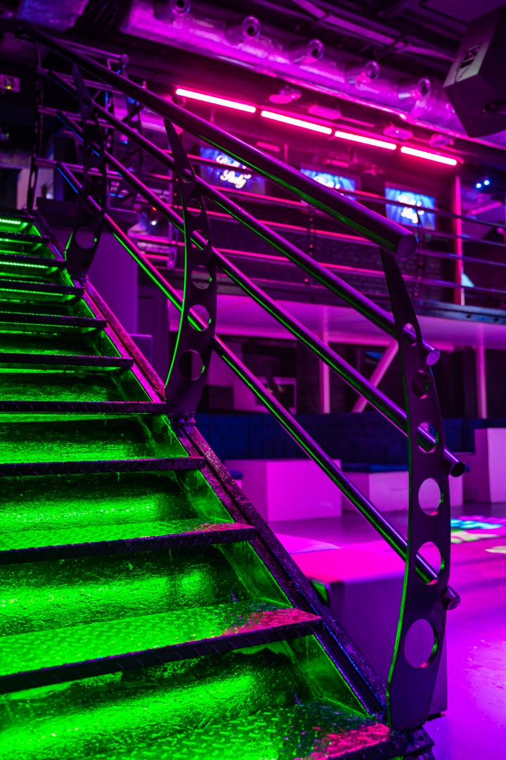 Metalowe schody z poręczą oświetlone neonowym, zielonym światłem, prowadzące na wyższy poziom w klubie z fioletowym oświetleniem.