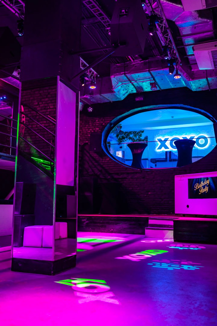 Wnętrze klubu nocnego z oświetleniem neonowym w odcieniach różu, fioletu i zieleni, widoczne schody, okrągłe okno z napisem 'XOXO' i dekoracje na stolikach w tle.