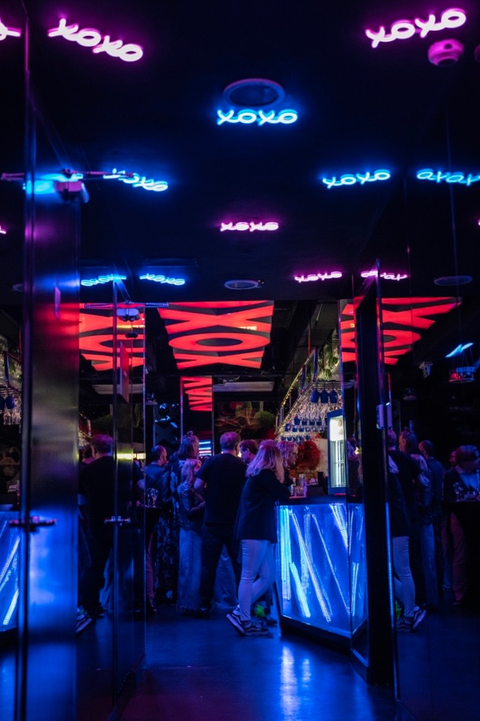 Wnętrze klubu nocnego z neonowymi napisami 'xoxo' na suficie i podświetlonym barem, wypełnione gośćmi podczas wydarzenia.