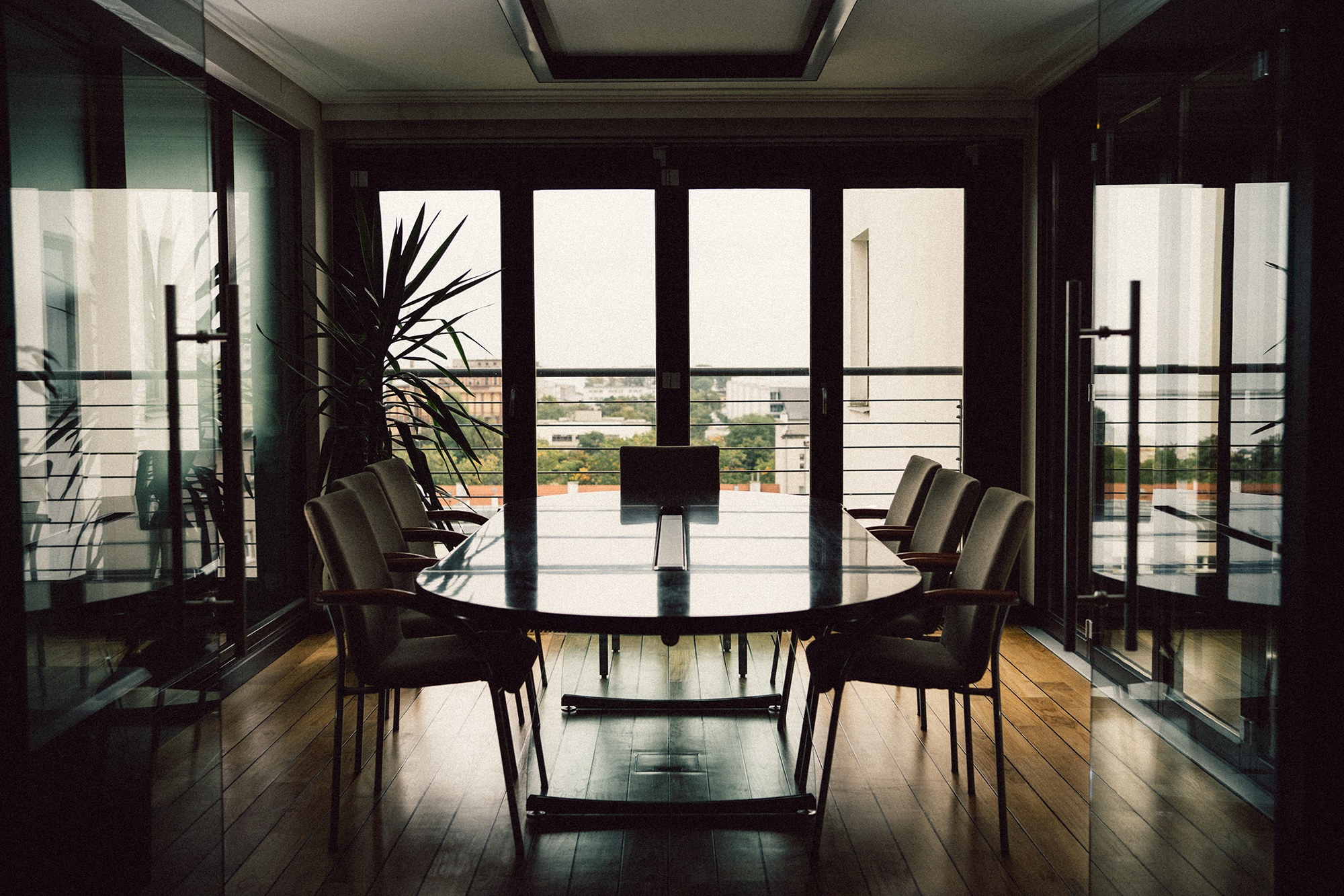 Sala konferencyjna z dużym stołem i krzesłami, widok na miasto przez okna. Minimalistyczny design, drewniana podłoga, roślina w rogu. Stonowana kolorystyka.