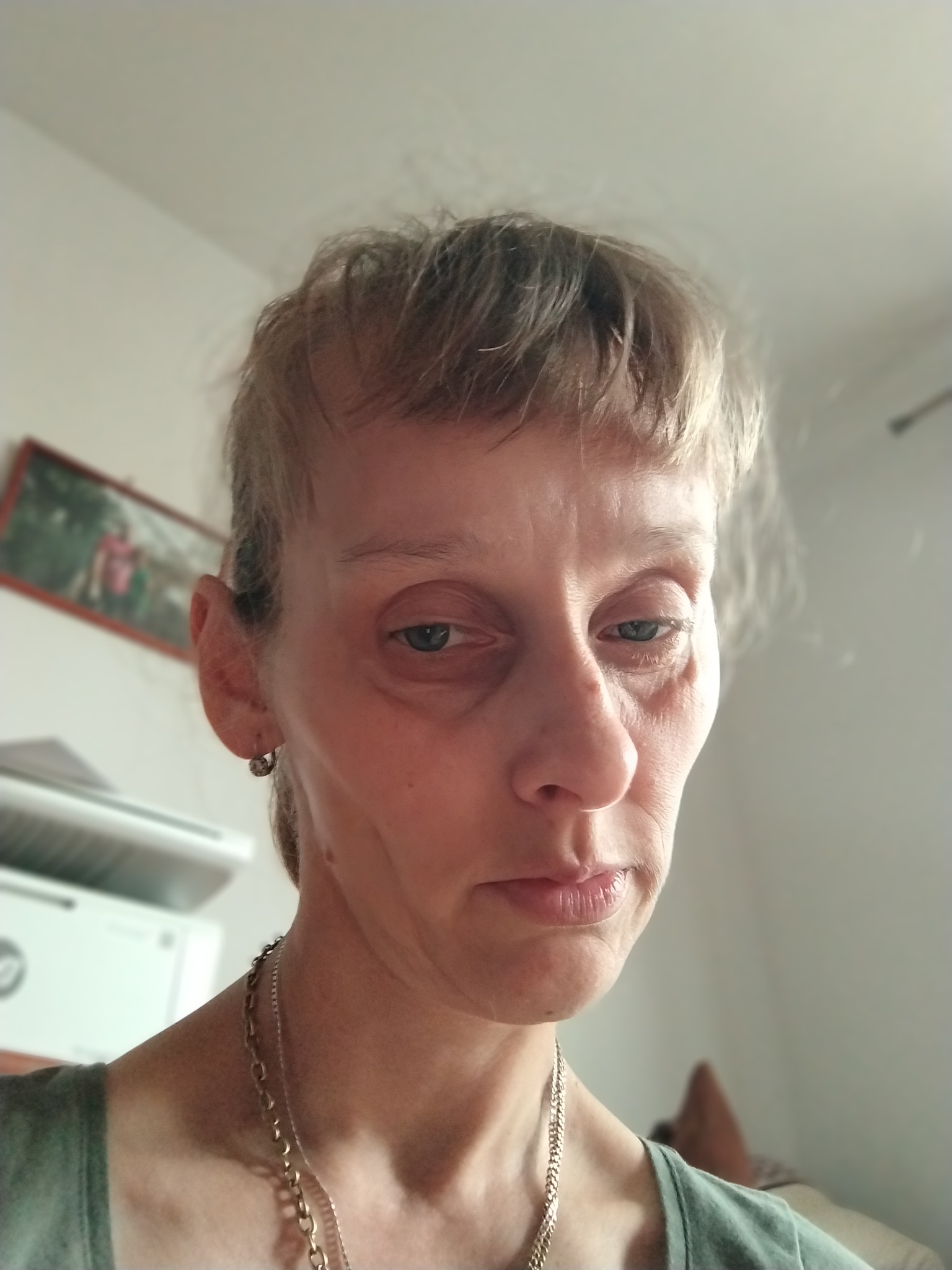 Portret kobiety z krótkimi blond włosami, srebrnym kolczykiem i dwoma łańcuszkami na szyi, w tle widoczny fragment ściany z obrazkiem w ramce i urządzeniem biurowym.