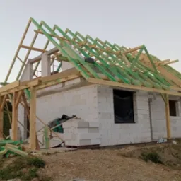 Budowa domu: ściany z białych bloczków, drewniana konstrukcja dachu z zielonymi elementami, widok na pagórkowaty krajobraz w tle.