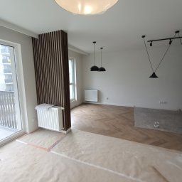 Oleksandr Khalo KHOM-BUD - Wnętrze w trakcie prac wykończeniowych: parkiet ułożony w jodełkę, osłonięty folią malarską, widoczne listwy dekoracyjne z ciemnego drewna, grzejniki, lampy wiszące oraz sufitowa lampa z abażurem...