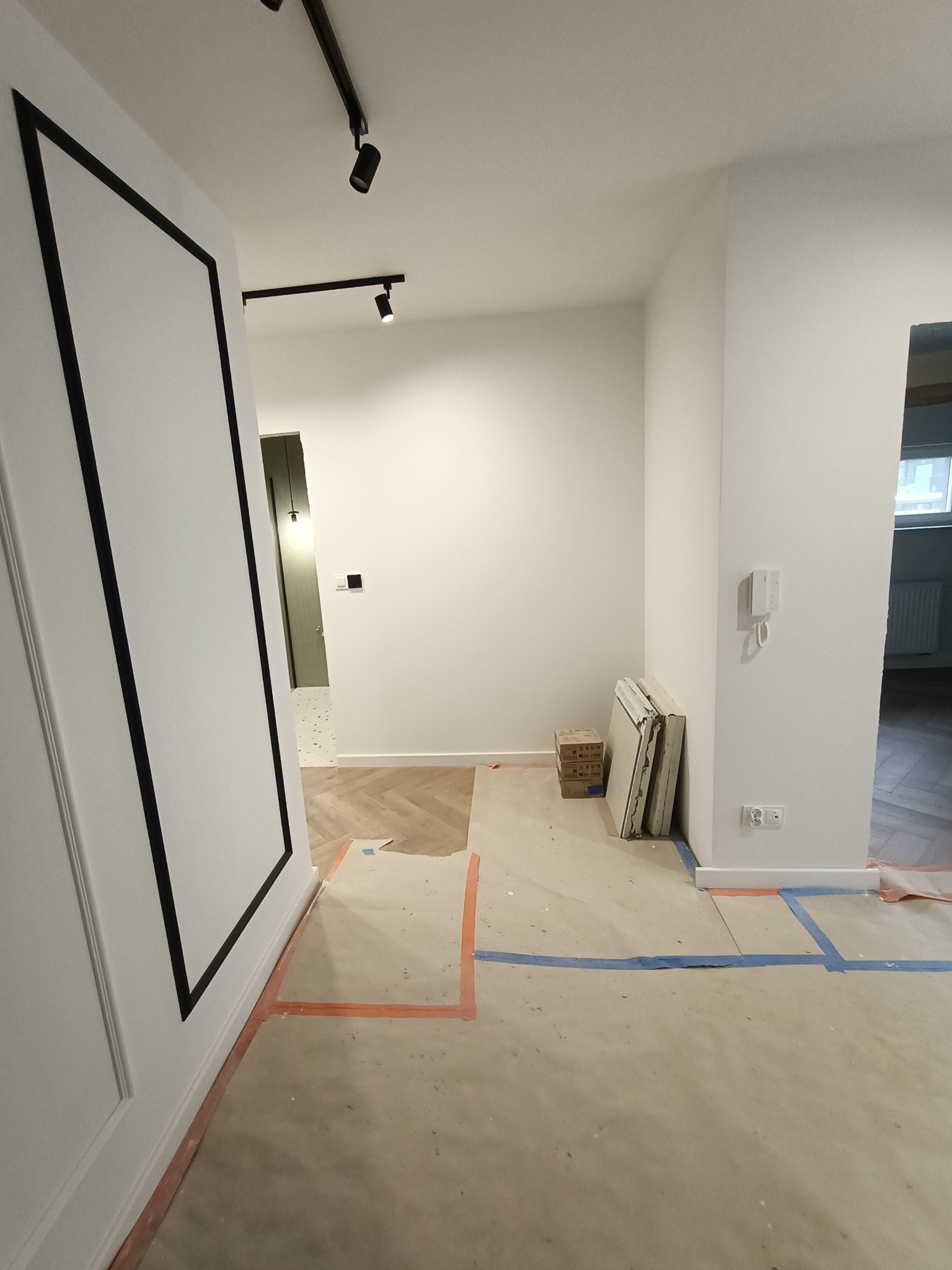 Korytarz w trakcie prac wykończeniowych: zabezpieczona podłoga, częściowo ułożony parkiet w jodełkę, białe ściany z czarnymi dekoracyjnymi listwami, reflektory sufitowe, płyty gipsowe oparte o ścianę.