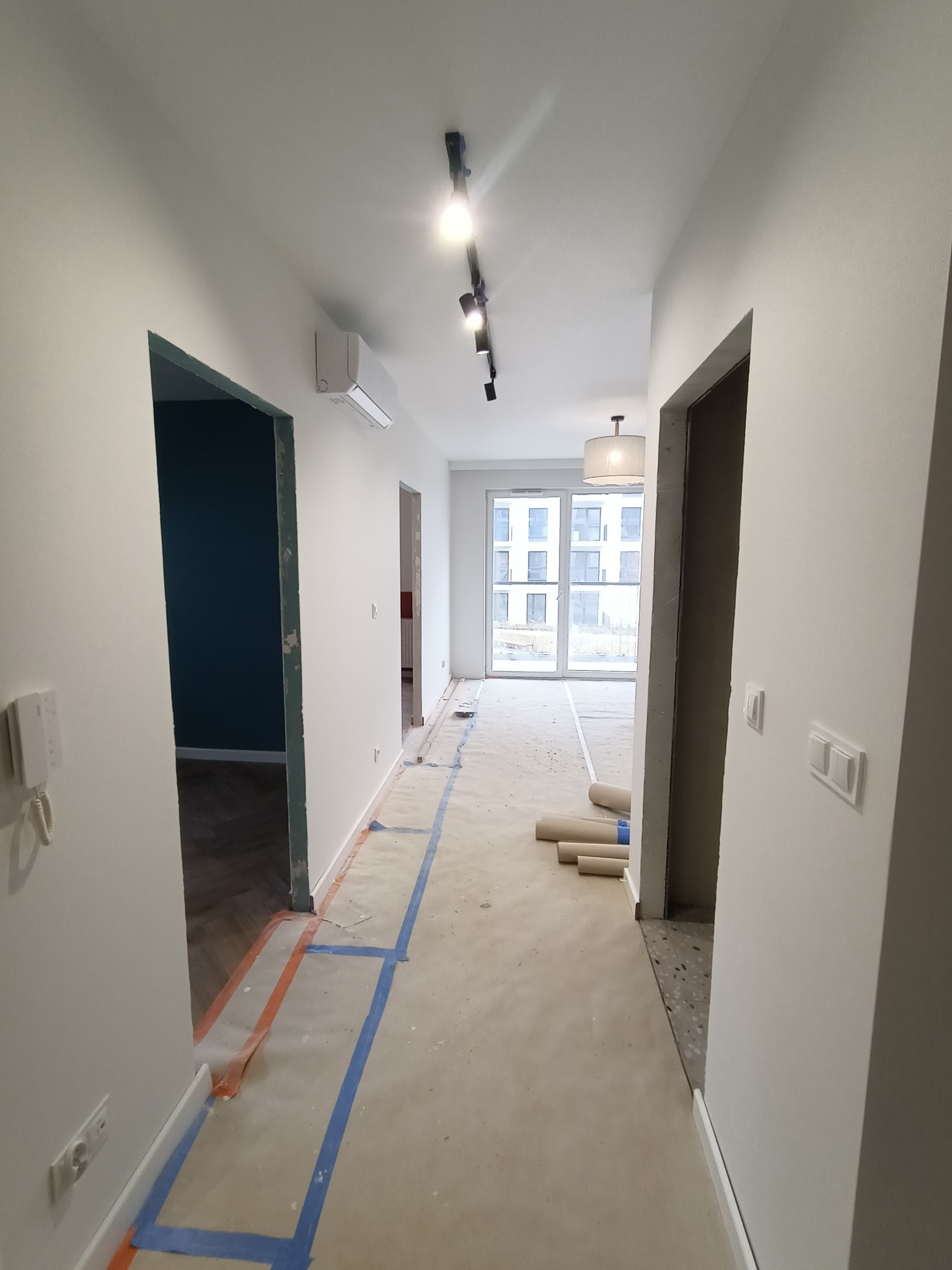 Korytarz w trakcie prac wykończeniowych: ściany pomalowane na biało, podłoga zabezpieczona folią i taśmą malarską, widoczne rolki materiału podłogowego, surowa ościeżnica drzwiowa i fragment...