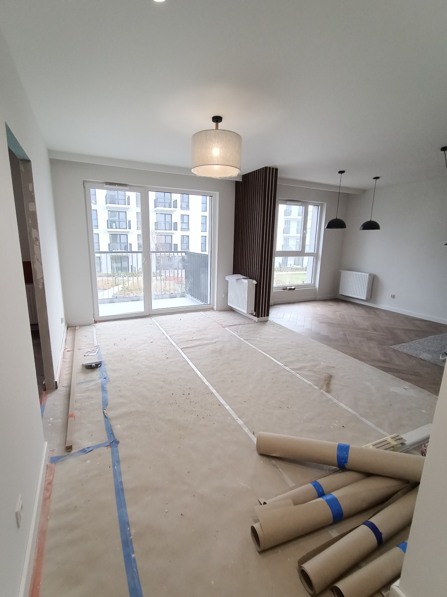 Wnętrze mieszkania w trakcie prac wykończeniowych: zabezpieczona folią podłoga, widoczne rolki papieru ochronnego z niebieską taśmą, częściowo ułożony parkiet w jodełkę, grzejniki, okno balkonowe...