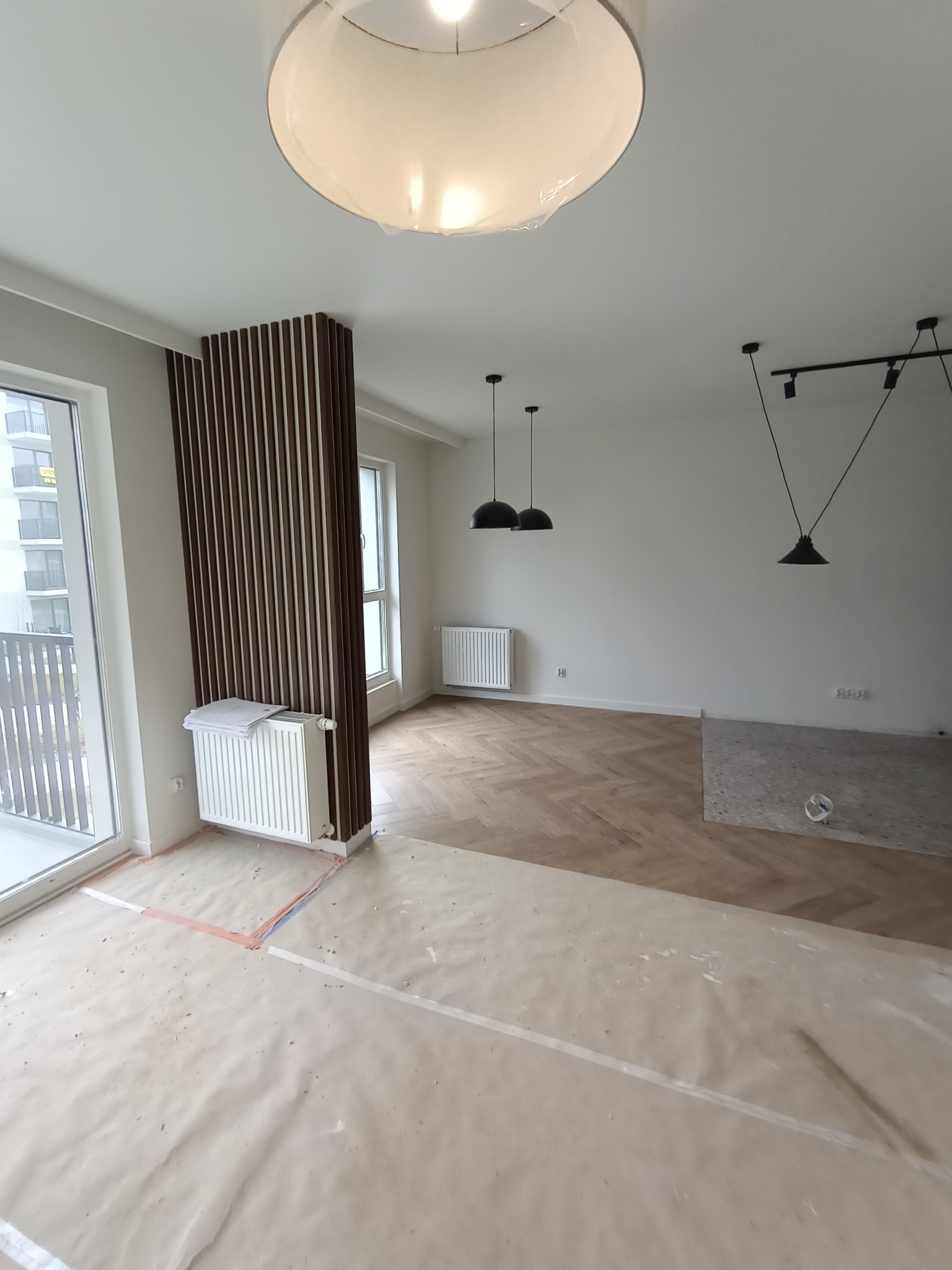 Wnętrze w trakcie prac wykończeniowych: parkiet ułożony w jodełkę, osłonięty folią malarską, widoczne listwy dekoracyjne z ciemnego drewna, grzejniki, lampy wiszące oraz sufitowa lampa z abażurem...