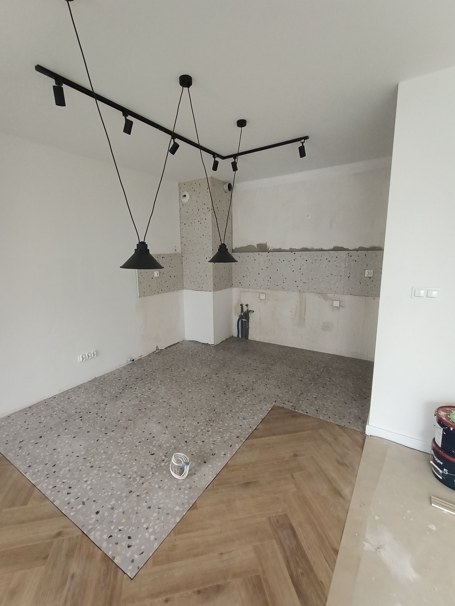 Wnętrze w trakcie remontu: Połączenie podłogi z lastryko i ułożonego parkietu w jodełkę, widoczne lampy sufitowe na szynach i puszki z farbą.