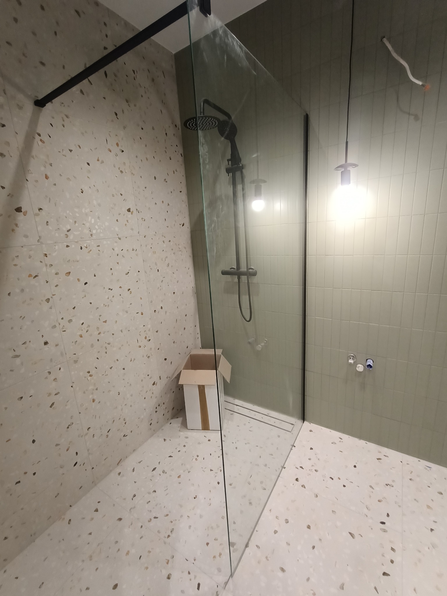 Nowoczesna kabina prysznicowa typu walk-in z czarną armaturą, szklaną ścianą i odpływem liniowym, na ścianach i podłodze płytki lastryko, widoczna kartonowa skrzynka w rogu.