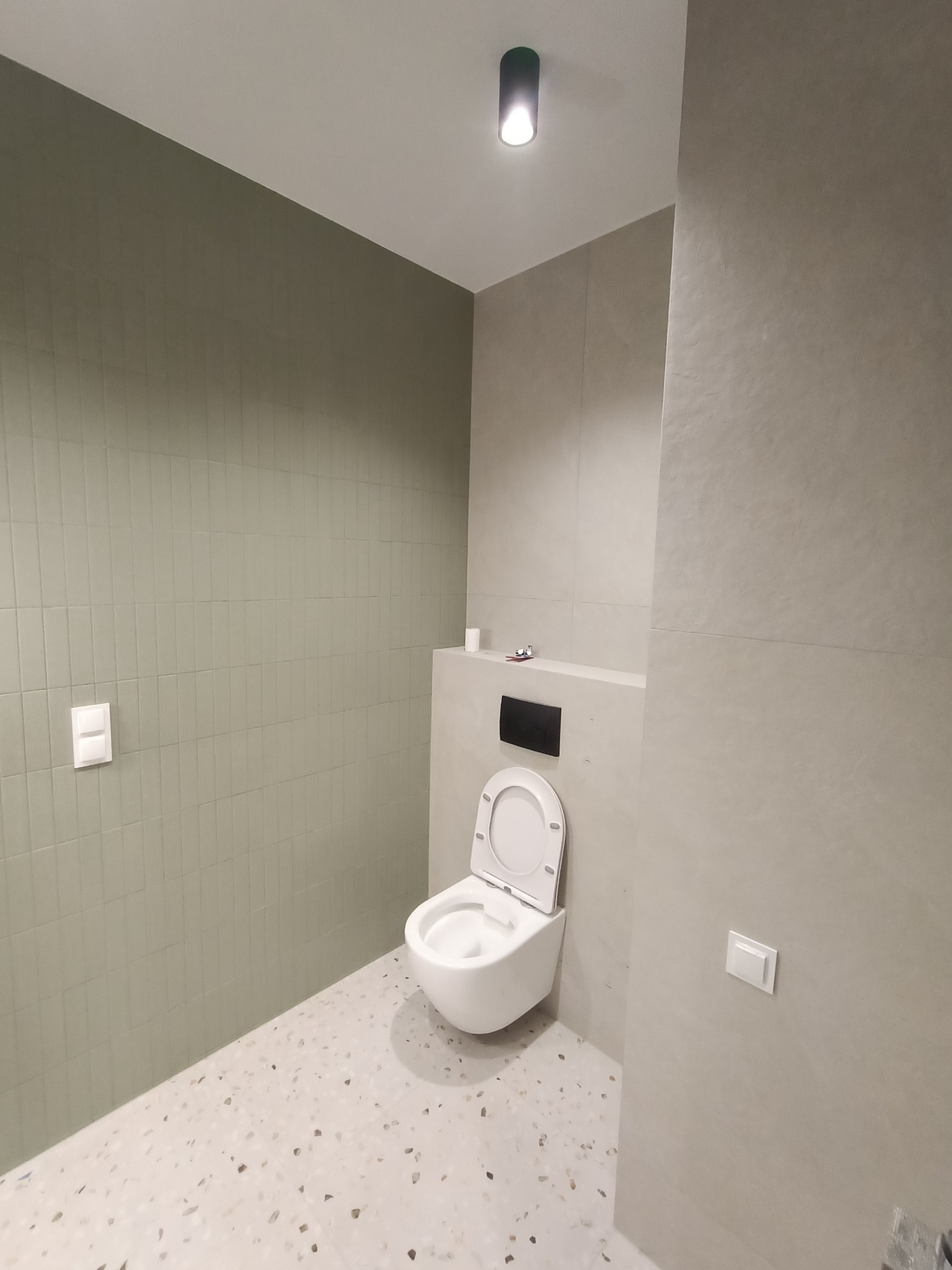 Nowoczesna łazienka z wiszącą toaletą, zielonymi płytkami na ścianie i podłogą w stylu lastryko, minimalistyczne oświetlenie sufitowe.