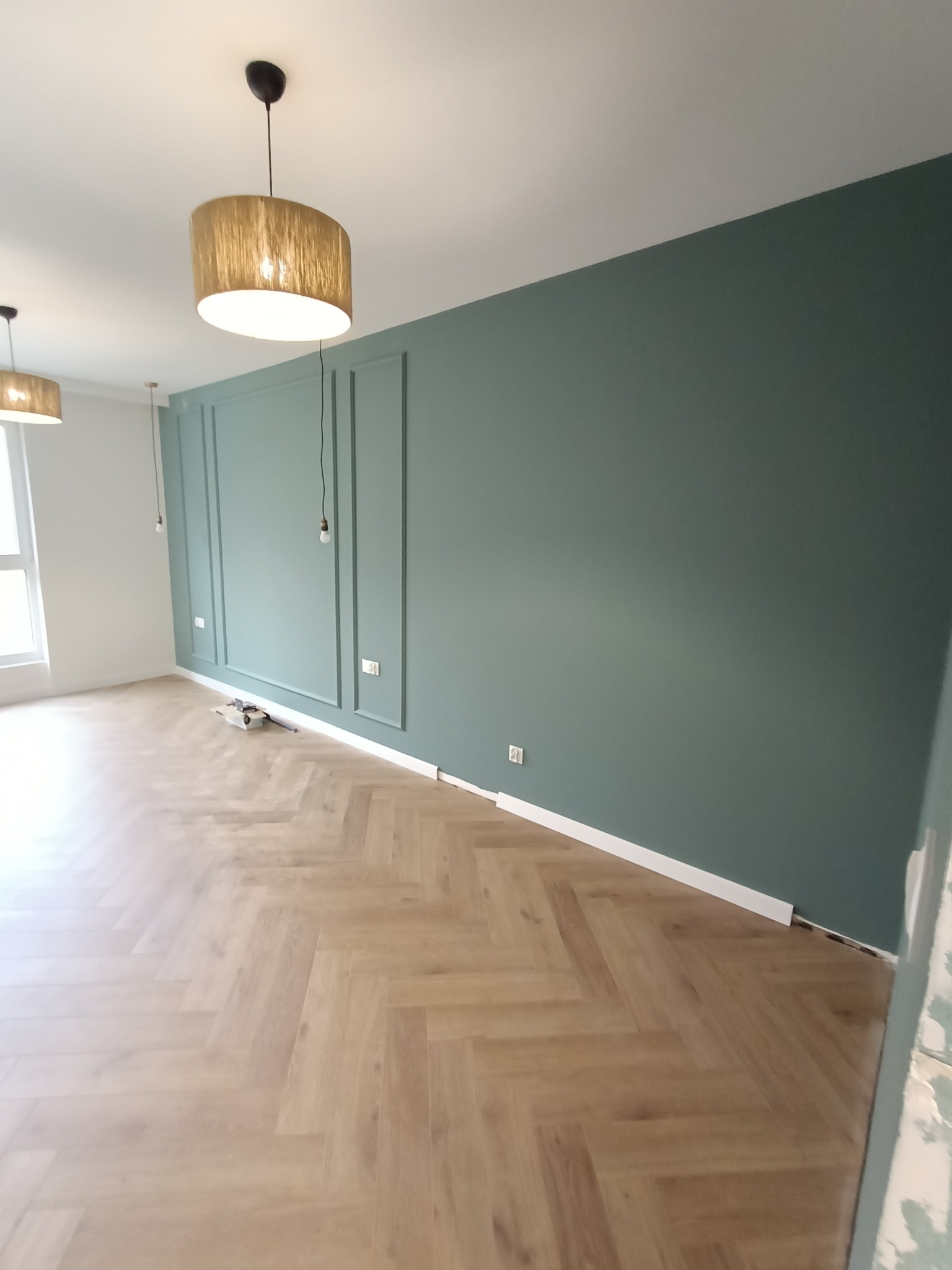 Wykończone wnętrze z podłogą ułożoną w jodełkę, pomalowaną na zielono ścianą z dekoracyjnymi listwami i wiszącymi lampami z abażurami