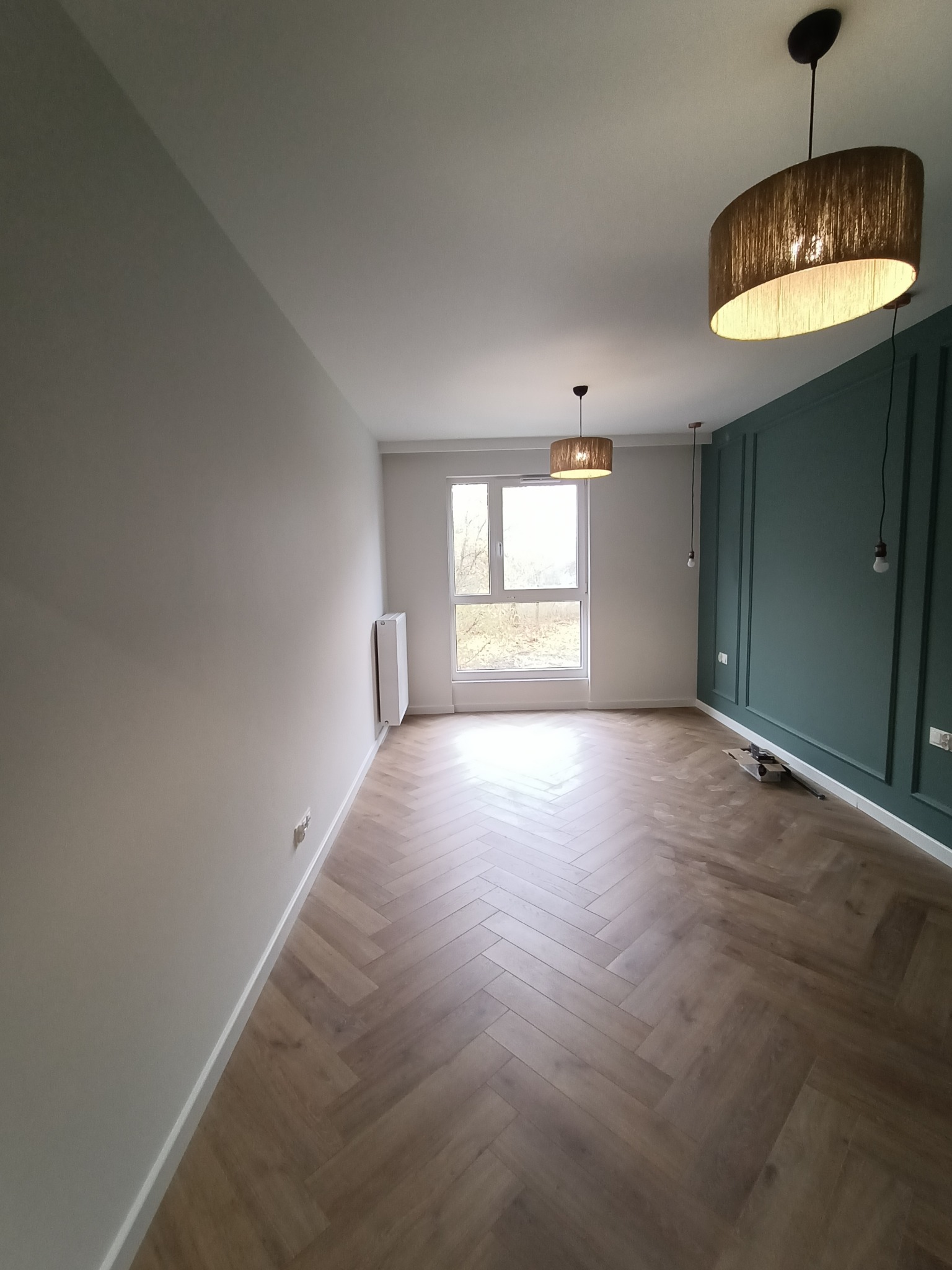 Salon po remoncie z parkietem ułożonym w jodełkę, ścianą pomalowaną na butelkową zieleń z dekoracyjnymi listwami i nowoczesnym oświetleniem.