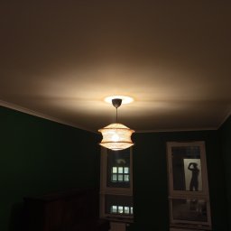 Fachman Damian Fryz - Włączona, pleciona lampa wisząca z żarówką, oświetlająca sufit w pokoju z zielonymi ścianami. W oknie widoczna sylwetka osoby robiącej zdjęcie.