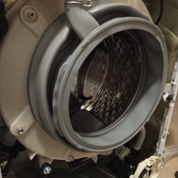 Fachman Damian Fryz - Rozłożona pralka z widocznym bębnem i elementami konstrukcyjnymi. Gumowy kołnierz uszczelniający bęben. Widoczne ślady demontażu, narzędzia w tle.