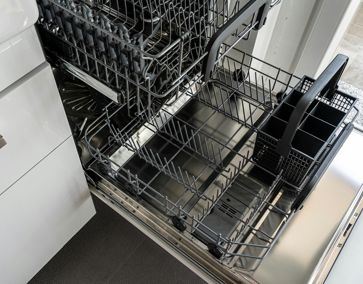 Otwarta, pusta zmywarka z wysuniętymi koszami w nowoczesnej kuchni. Widoczne metalowe kosze na naczynia i sztućce, wnętrze ze stali nierdzewnej.