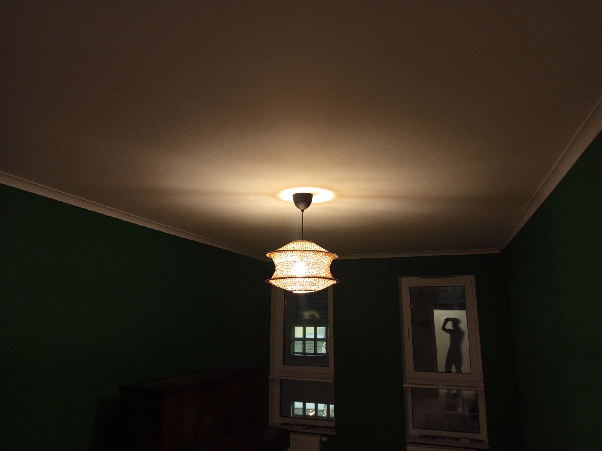 Włączona, pleciona lampa wisząca z żarówką, oświetlająca sufit w pokoju z zielonymi ścianami. W oknie widoczna sylwetka osoby robiącej zdjęcie.