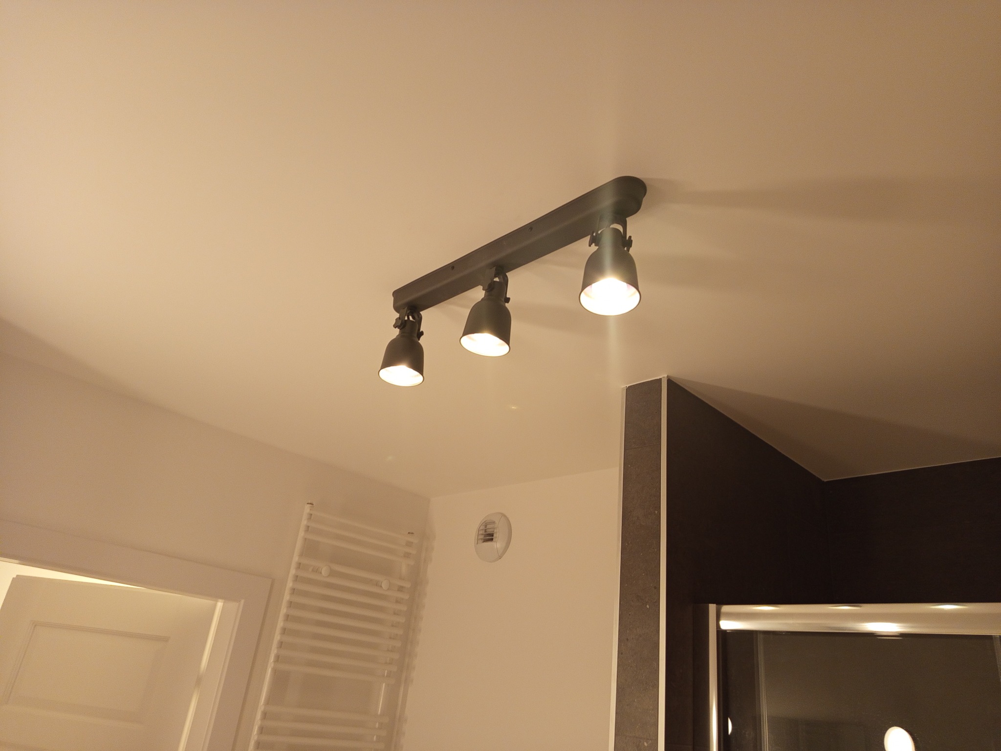 Zamontowana listwa z trzema reflektorami sufitowymi w łazience, oświetlając wnętrze. Widoczny fragment grzejnika drabinkowego i fragment kabiny prysznicowej.