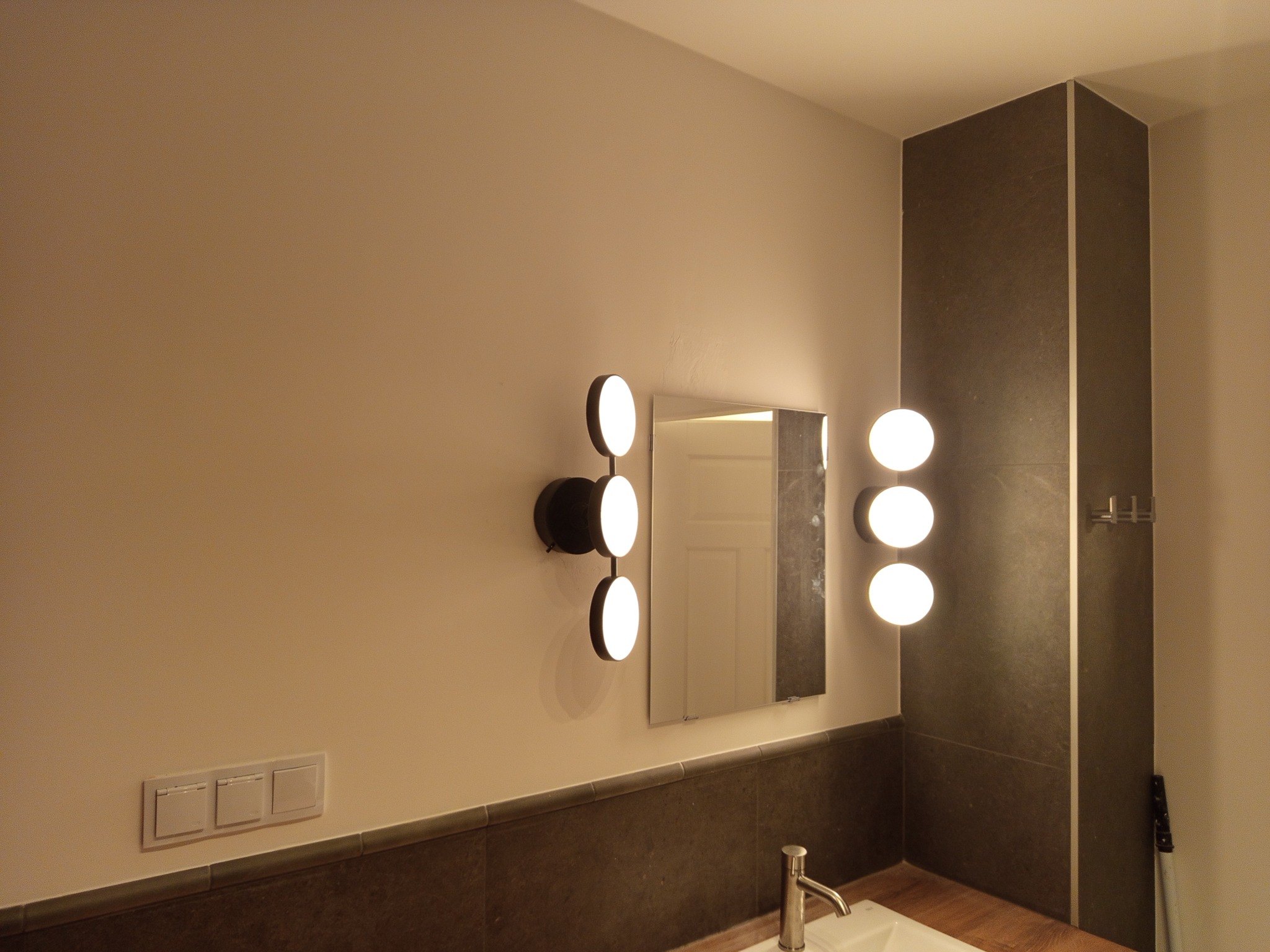 Nowoczesna łazienka z podświetlanym lustrem i designerskimi lampami kulistymi po bokach. Umywalka z baterią i ciemna glazura dopełniają wystrój.