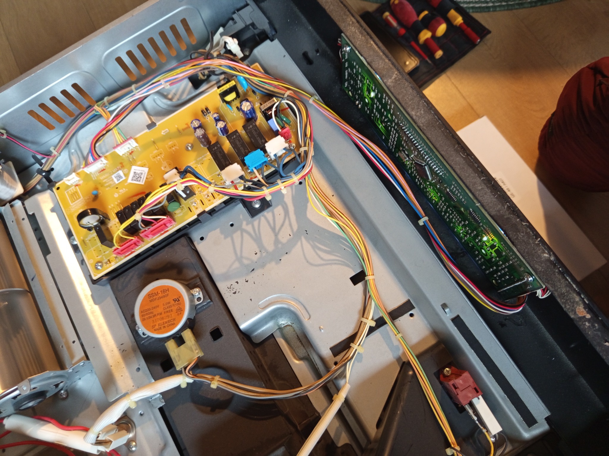 Wnętrze piekarnika Combi Samsung z widocznym modułem sterującym i wyświetlaczem po wymianie. Żółta płytka drukowana z kolorowymi kablami. Narzędzia w tle.