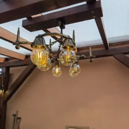 Kuta, metalowa lampa wisząca z żarówkami Edison na tle drewnianej konstrukcji zadaszenia tarasu i beżowej ściany.
