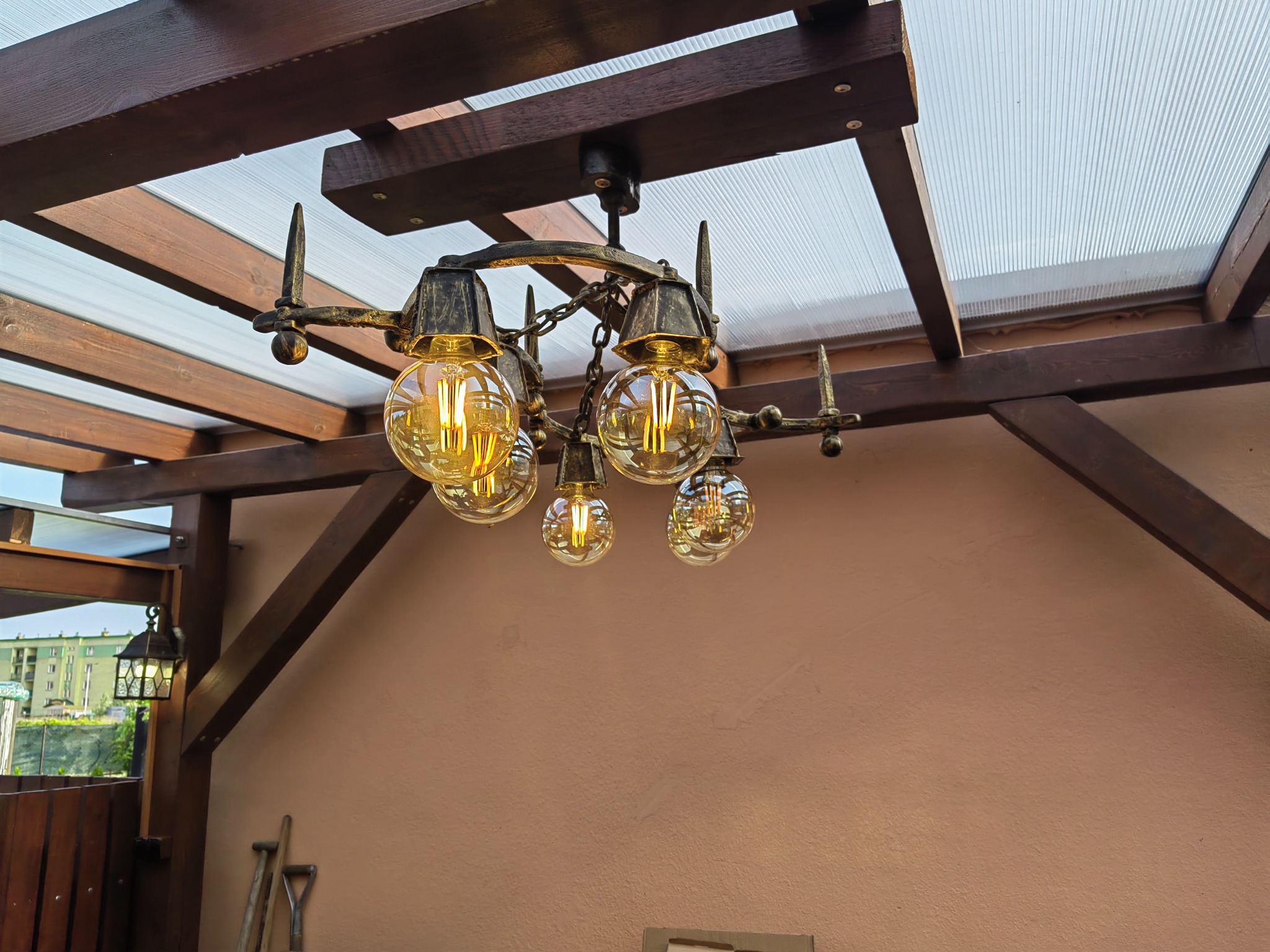 Kuta, metalowa lampa wisząca z żarówkami Edison na tle drewnianej konstrukcji zadaszenia tarasu i beżowej ściany.