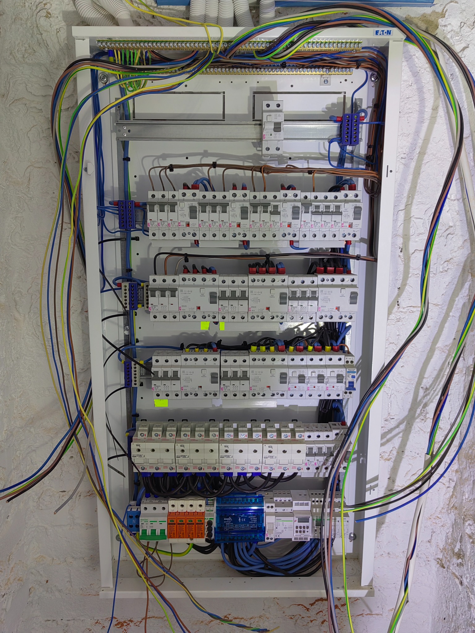Rozdzielnica elektryczna firmy z Pisza, z licznymi zabezpieczeniami i przewodami o różnych kolorach, zamontowana na ścianie o nierównej fakturze.