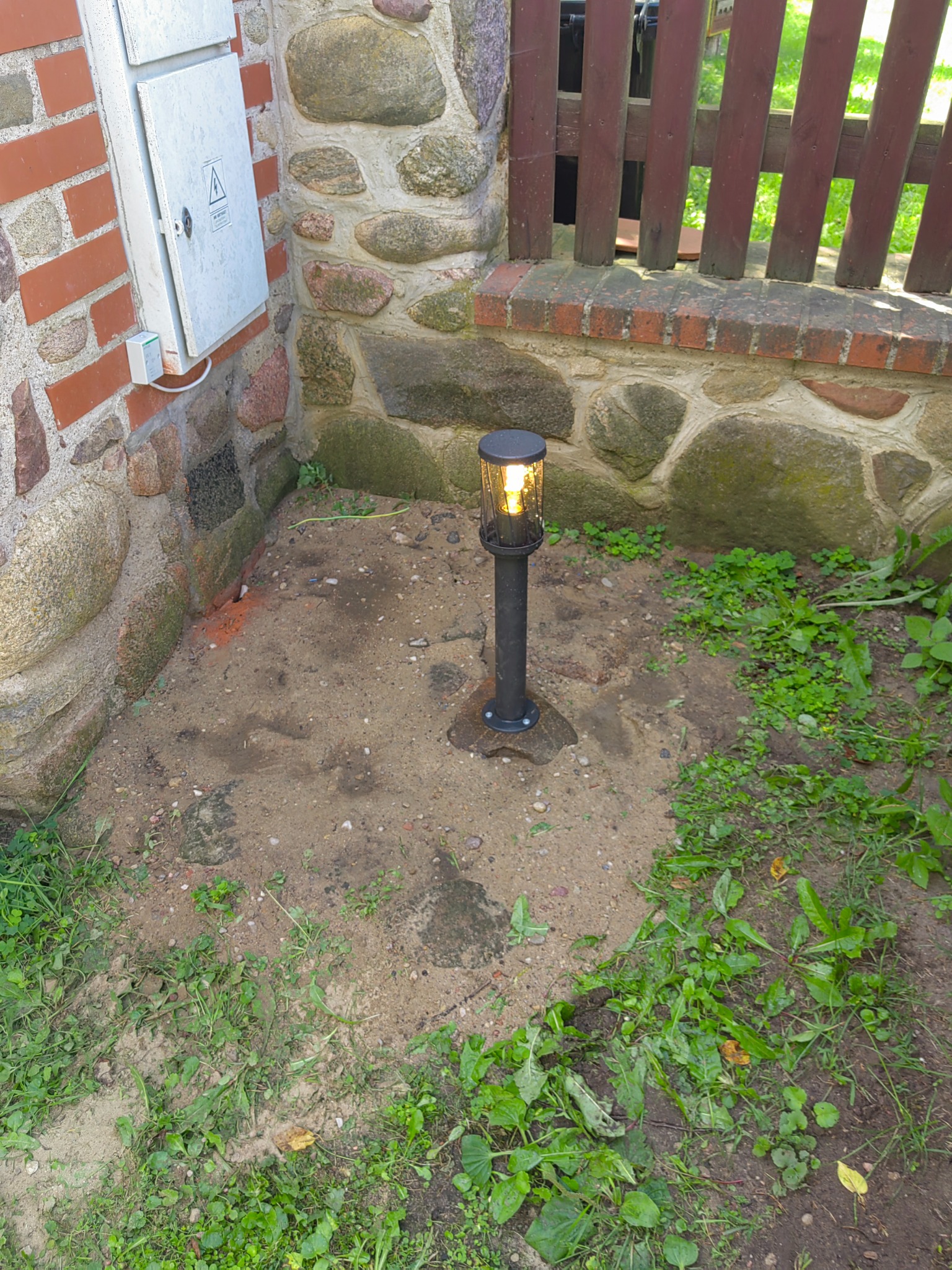 Zamontowana lampa ogrodowa w czarnej obudowie, świecąca ciepłym światłem, umieszczona na piaszczystym podłożu przy kamienno-ceglanym murze, obok skrzynki elektrycznej i drewnianego płotu.