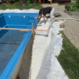 Montaż prefabrykowanego basenu w ogrodzie: pracownik wyrównuje podłoże z białego kruszywa wokół niebieskiej niecki basenowej z widocznym zbrojeniem i drewnianymi stemplami stabilizującymi.