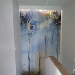 Ściana w klatce schodowej z fototapetą przedstawiającą akwarelowy las w odcieniach błękitu i brązu, podświetlona od góry, z fragmentem schodów z drewnianą poręczą.