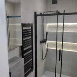 Nowoczesna łazienka z kabiną prysznicową w czarnej ramie, grzejnikiem drabinkowym w kolorze czarnym, umywalką z szarego betonu architektonicznego i podświetlanymi płytkami dekoracyjnymi na ścianie.