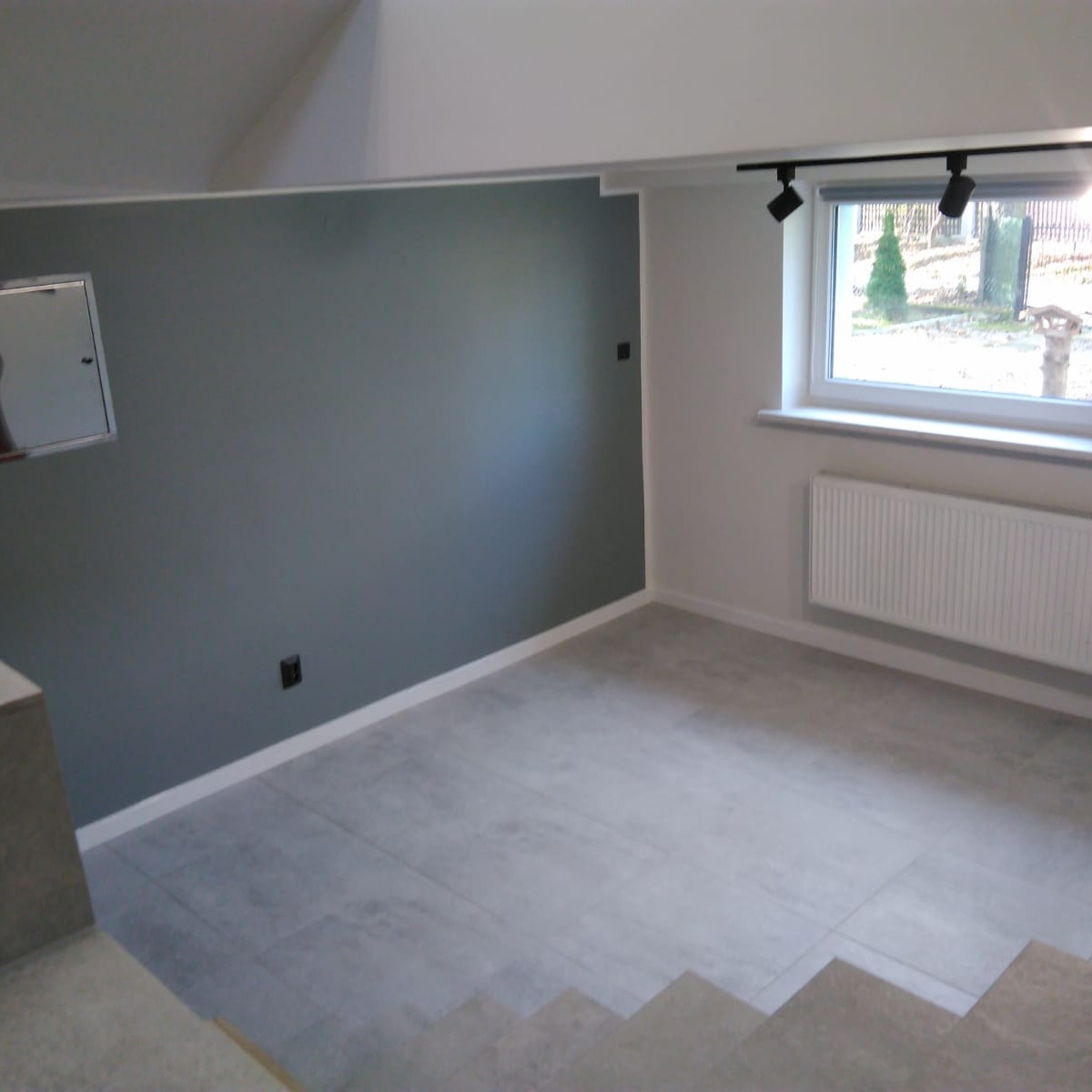 Pomieszczenie po remoncie z szarą ścianą, szarą podłogą z płytek, białym grzejnikiem pod oknem i czarnymi reflektorami sufitowymi.