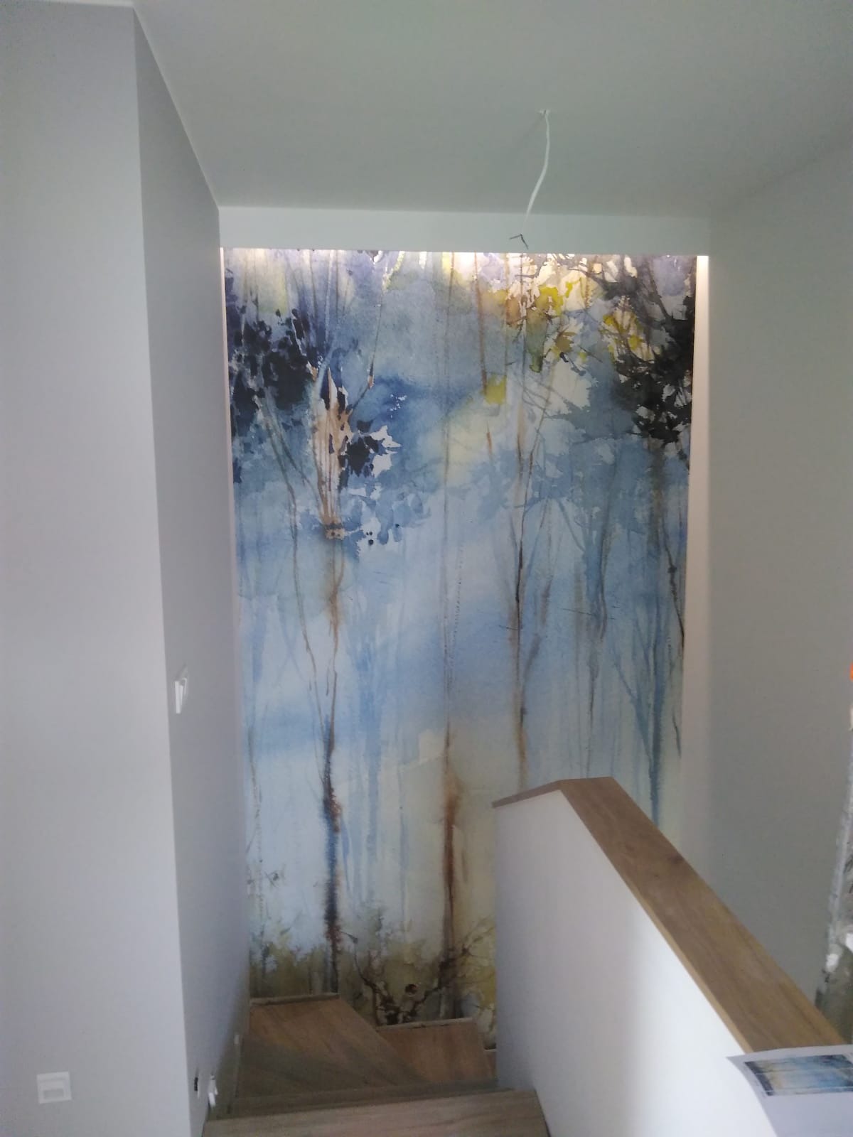 Ściana w klatce schodowej z fototapetą przedstawiającą akwarelowy las w odcieniach błękitu i brązu, podświetlona od góry, z fragmentem schodów z drewnianą poręczą.