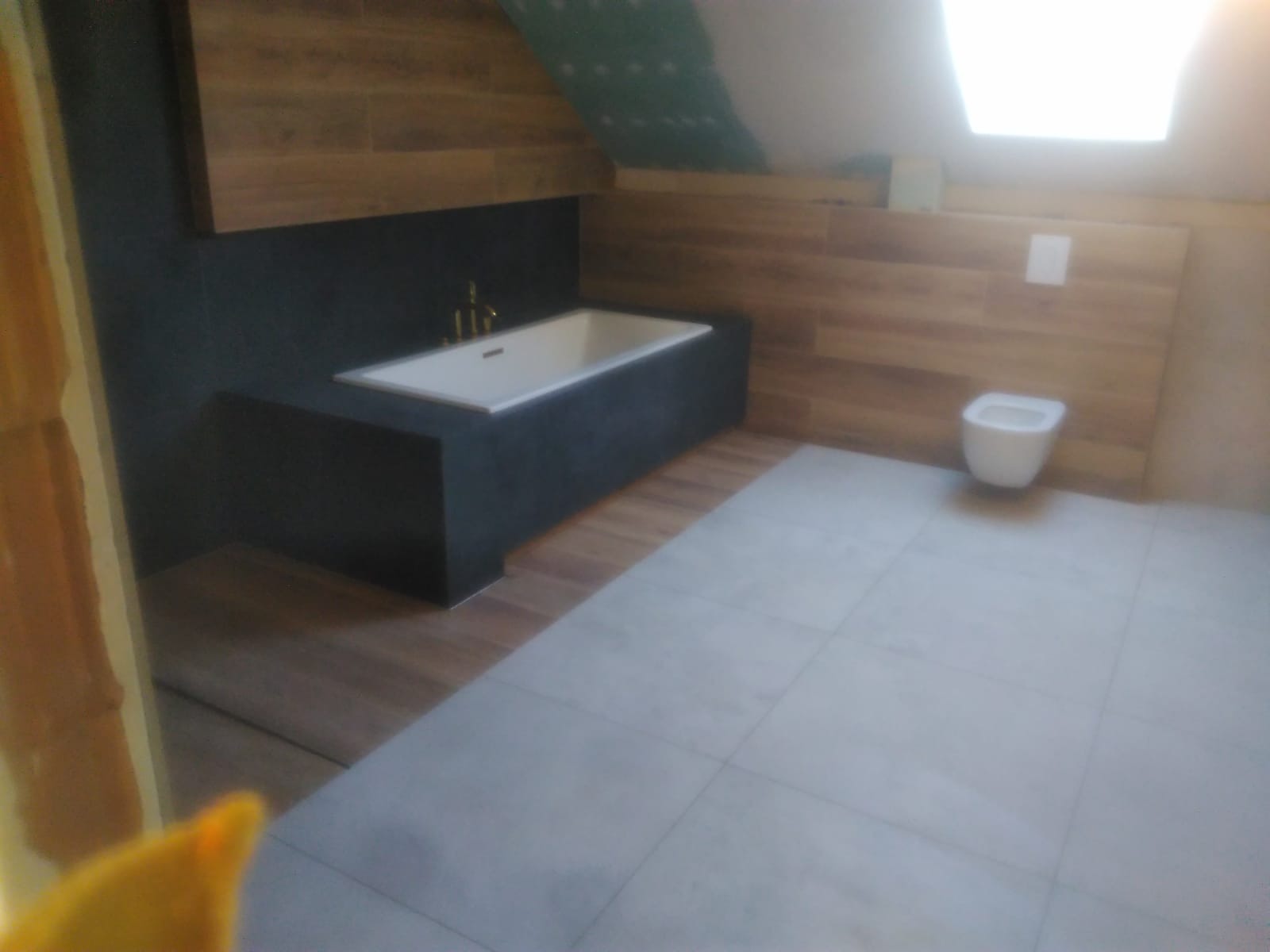 Wykończona łazienka na poddaszu z wanną w zabudowie z ciemnej płyty, drewnopodobnymi płytkami na ścianach i szarą podłogą z płytek, widoczna toaleta podwieszana.