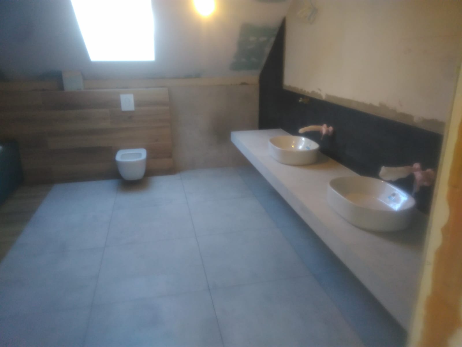 Łazienka na poddaszu z dwiema umywalkami na długim blacie, drewnopodobną okładziną ściany za toaletą i szarą glazurą podłogową.