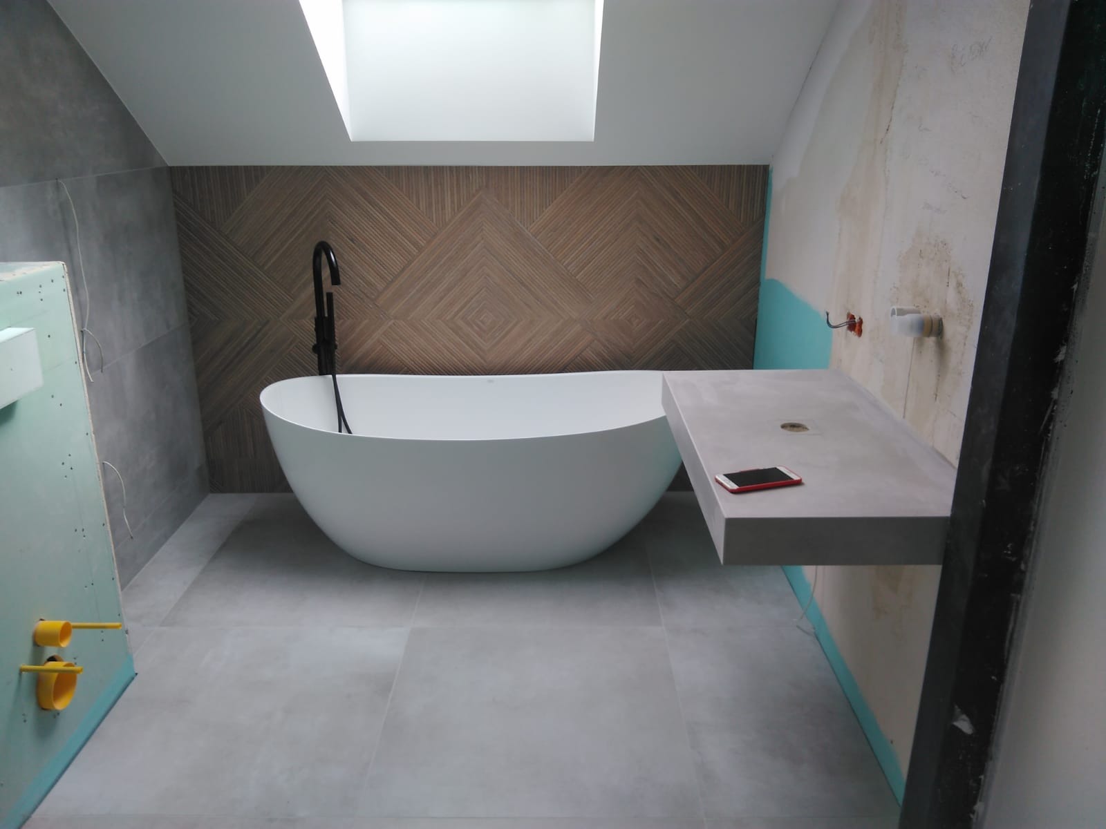 Wanna wolnostojąca z czarną armaturą na tle ściany z drewnopodobnych paneli, obok betonowy blat z leżącym smartfonem, sufit z oknem dachowym, widoczne elementy instalacji w trakcie prac...