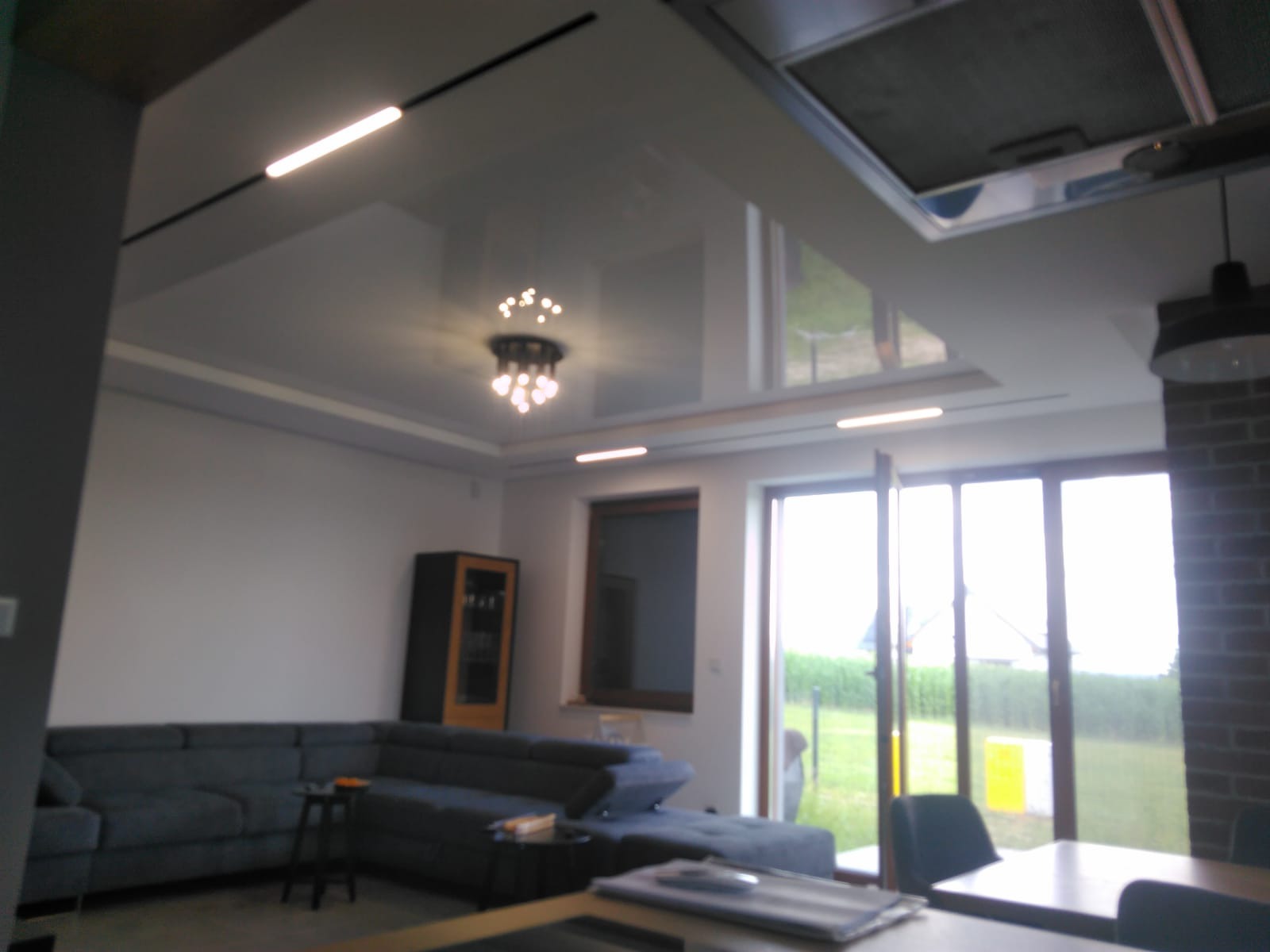 Nowoczesny salon z szarym narożnikiem, błyszczącym sufitem napinanym z oświetleniem LED i żyrandolem, widok na ogród przez duże okna.