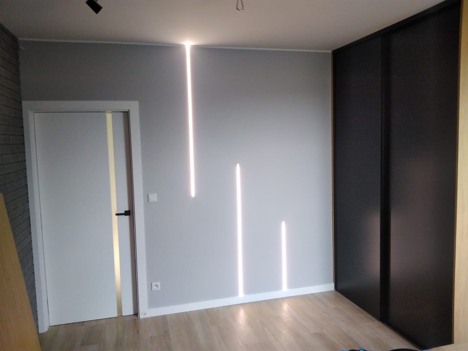 Szara ściana z trzema pionowymi wpuszczonymi liniami świetlnymi, białe drzwi z czarną klamką po lewej, podłoga z paneli, ciemna szafa przesuwna po prawej.