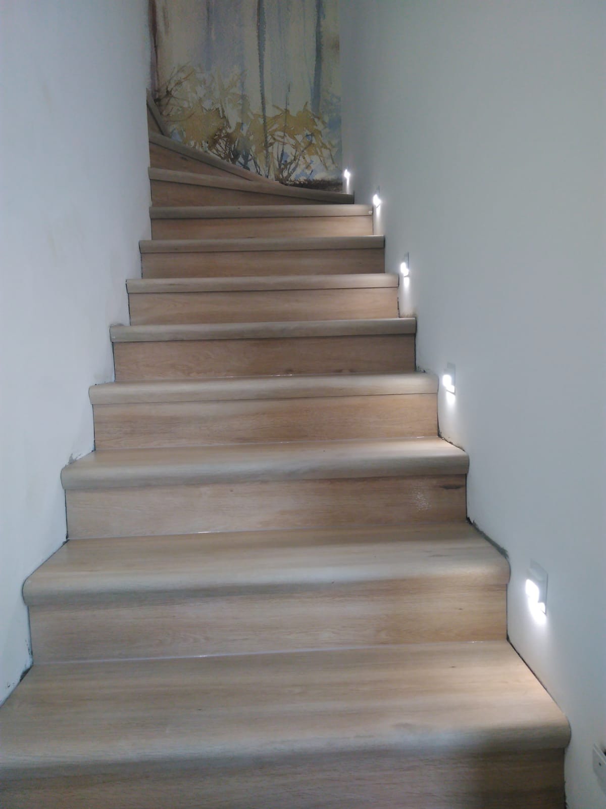 Drewniane schody z subtelnym oświetleniem punktowym w ścianie, prowadzące w górę, z abstrakcyjnym malowidłem na ścianie w tle.