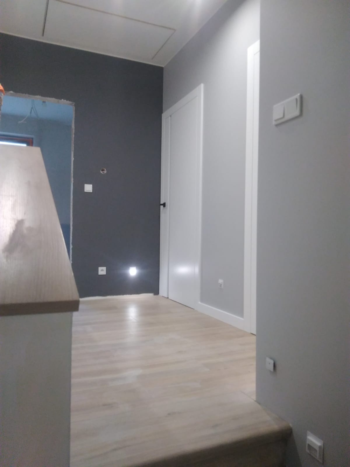 Korytarz po remoncie: szara ściana z wbudowanym oświetleniem punktowym, białe drzwi z czarną klamką, panele podłogowe imitujące drewno, otwór drzwiowy bez ościeżnicy w tle.